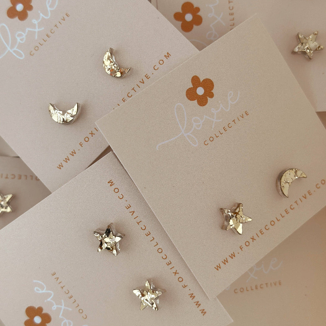 Gold celestial stud earrings, including stars, moons, and a crescent moon, displayed on beige cards from Foxie Collective.
