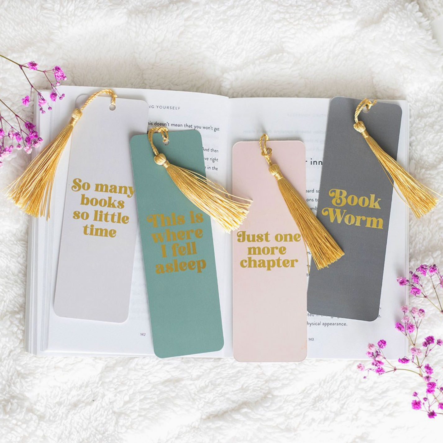 Four bookmarks with tassels on an open book, surrounded by flowers on a white surface.