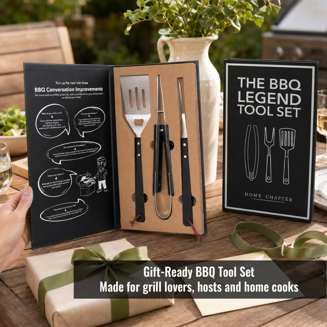 A gift-ready BBQ tool set displayed open on a wooden table, with its black-and-white booklet insert and cover standing beside it. A hand is visible holding the left edge of the insert, and the scene includes a wrapped gift box, green ribbon, plants, and dining table setting.