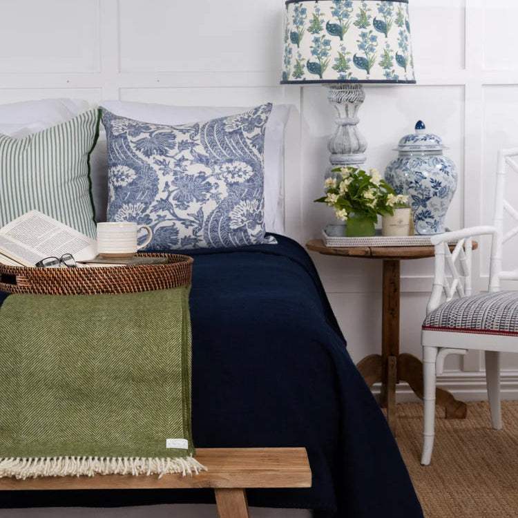 A styled bedroom scene featuring a navy blue throw blanket draped over a bed with blue and white patterned pillows, alongside a green woven throw blanket on a wooden bench, a blue and white ceramic vase, and a matching lamp.