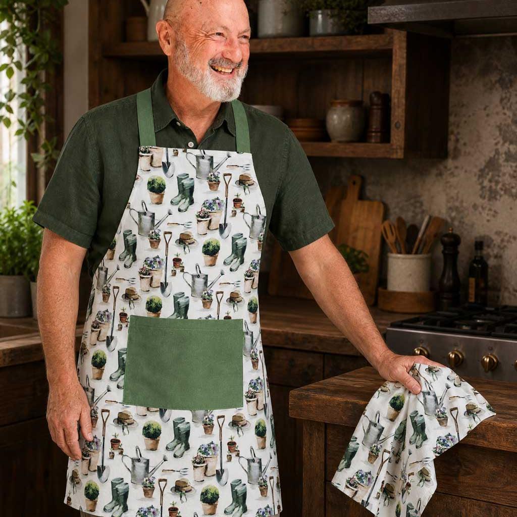 A smiling older man wearing a white apron with a green garden-themed print stands in a rustic kitchen. The apron has a green pocket and matching green straps, while he holds a matching kitchen towel with the same pattern.