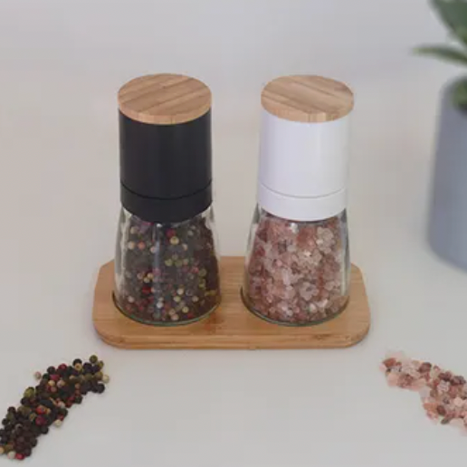 Two pepper grinders on a wooden stand with scattered peppercorns on a white surface.