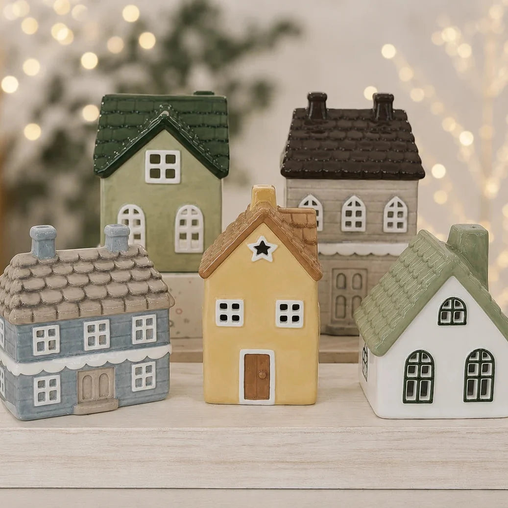A set of five miniature ceramic houses in various colors, arranged on a light wooden surface with a soft, blurred background of warm bokeh lights.