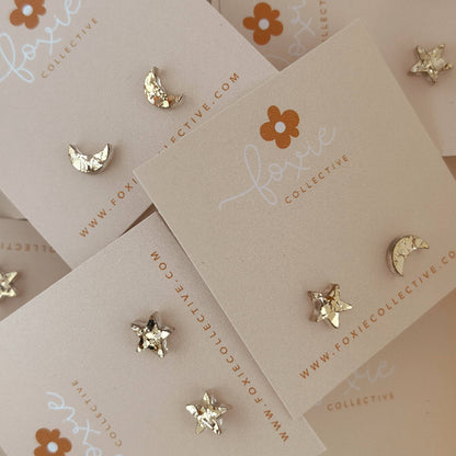 Gold celestial stud earrings, including stars, moons, and a crescent moon, displayed on beige cards from Foxie Collective.