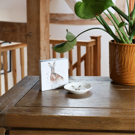A square art print or card featuring a watercolor rabbit in a meadow, styled on a rustic wooden table next to a small white dish and a potted green plant.