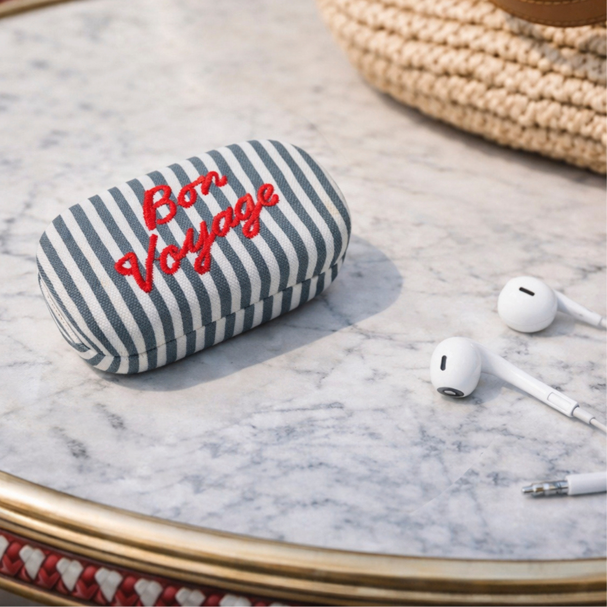 A striped fabric earbud case with red embroidered 'Bon Voyage' text, shown on a marble surface with white wired earbuds nearby.
