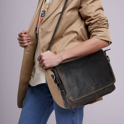 Person wearing a beige jacket and blue jeans holding a black leather bag against a gray background