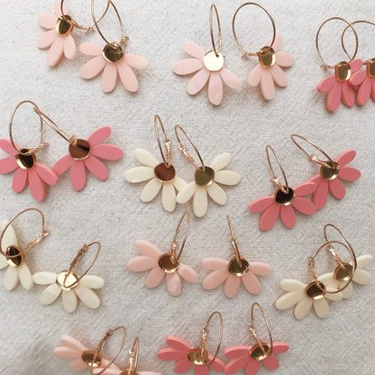 A flat-lay arrangement of daisy-shaped hoop earrings in pink, cream, and white, featuring gold-tone metal hoops and flower centers on a textured light gray fabric background.