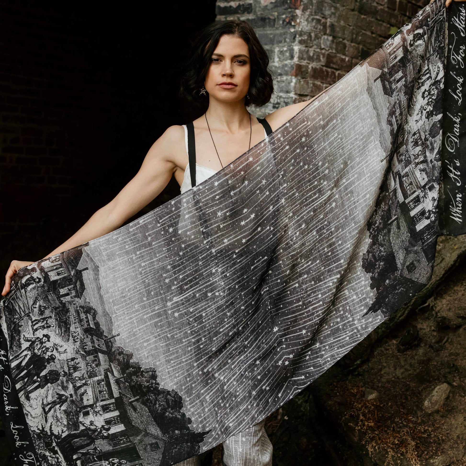 Woman holding a large black and white patterned fabric against a dark brick wall.
