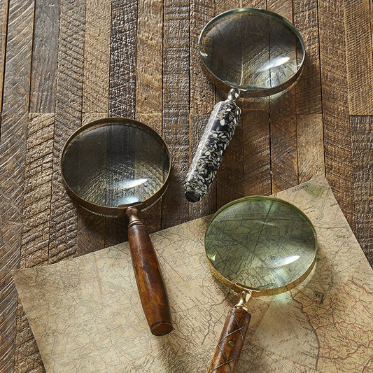 Three vintage-style magnifying glasses with wooden handles and brass rims are arranged on a rustic wood surface over an old map. Two have dark wood handles and one has a marbled gray-and-white handle.