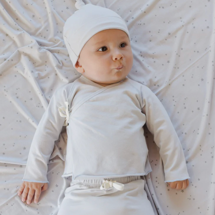 Baby lying on a textured white blanket wearing a white outfit and hat.