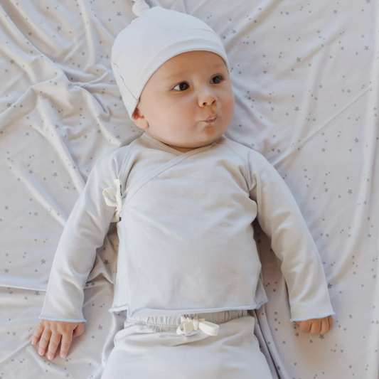 Baby lying on a textured white blanket wearing a white outfit and hat.