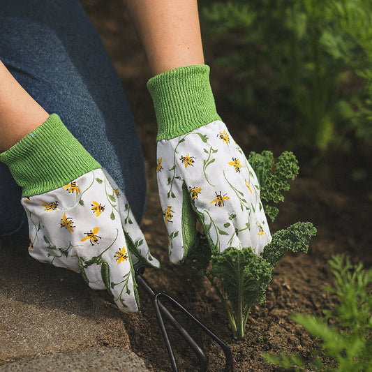 A close-up of a person gardening, wearing white gardening gloves with a green cuff and a yellow daisy floral print, planting kale in a garden bed.