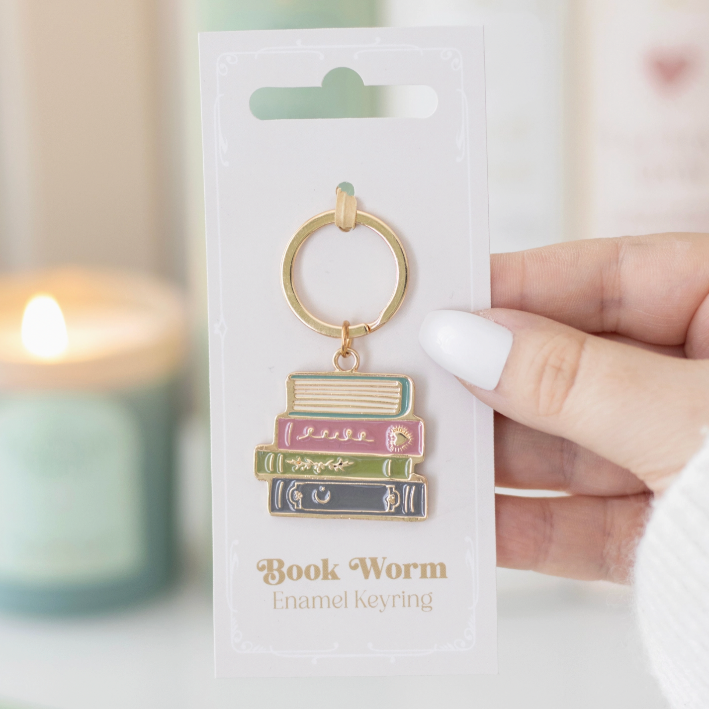 Book-themed keychain on a white card with 'Book Worm' text, held by a hand against a blurred candle background.