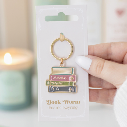 Book-themed keychain on a white card with 'Book Worm' text, held by a hand against a blurred candle background.