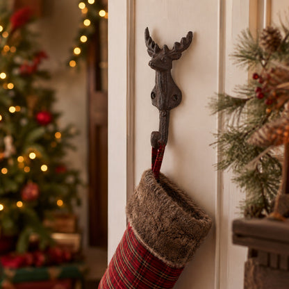 A red and green plaid Christmas stocking with a brown faux-fur cuff hangs from a dark metal reindeer wall hook on a white door, with a blurred decorated Christmas tree and holiday decor in the background.