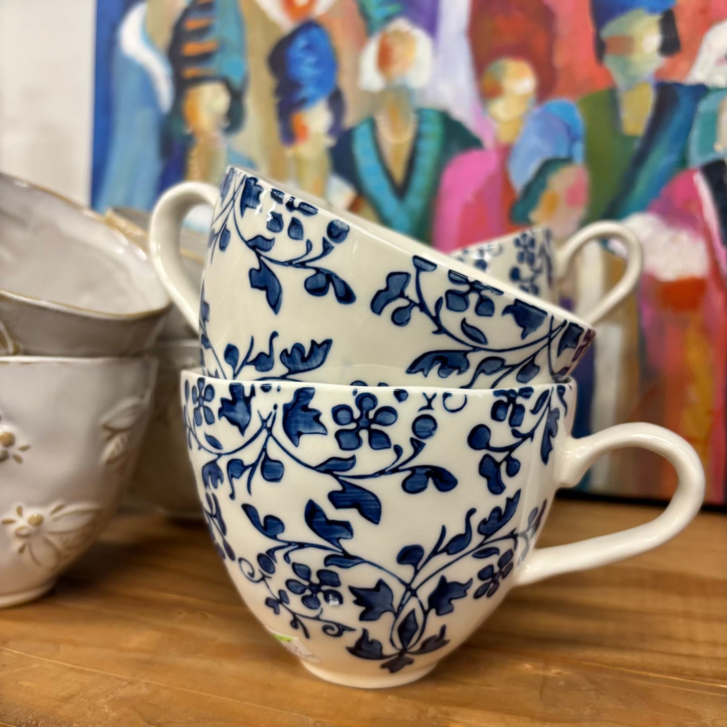 Two blue and white floral-patterned mugs stacked on a wooden surface with a colorful painting in the background.