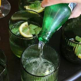 Green bottle being poured into a glass with lemon slices and ice cubes.