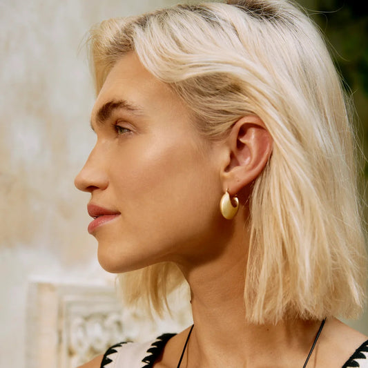 A close-up side profile of a woman with short platinum blonde hair, wearing a white sleeveless top with a black scalloped neckline and gold hoop earrings.
