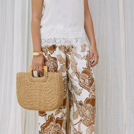 A person wearing wide-leg white pants with a brown floral print, holding a woven straw tote bag, standing in front of white curtains.