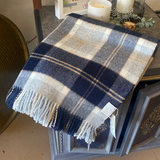 A folded silver and blue checkered wool throw blanket with fringes displayed on a grey stand, with a candle and decorative items in the background.
