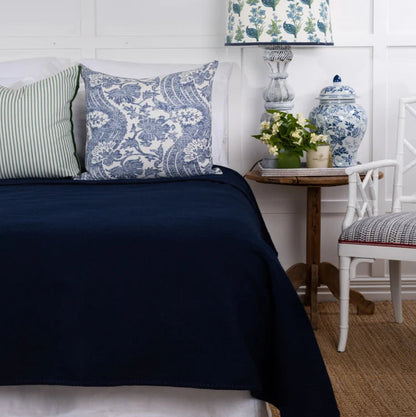 A styled bedroom corner featuring a bed with a dark navy blue blanket, a blue and white damask patterned pillow, and a green and white striped pillow. A matching blue and white patterned table lamp sits on a small wooden side table next to a white chair.