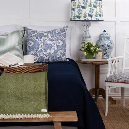 A styled bedroom scene featuring a navy blue throw blanket draped over a bed with blue and white patterned pillows, alongside a green woven throw blanket on a wooden bench, a blue and white ceramic vase, and a matching lamp.