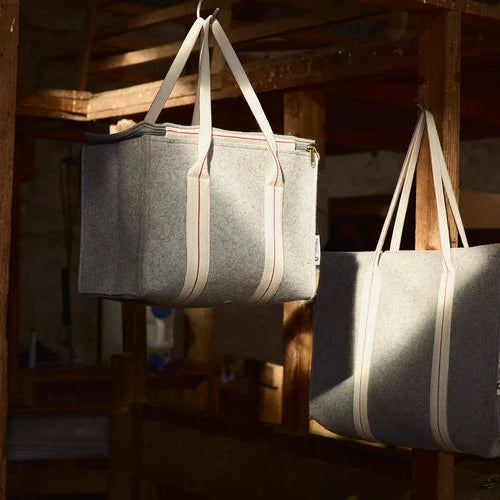 Two gray felt tote bags with cream-colored handles hang from metal hooks in a rustic wooden room with dramatic side lighting.