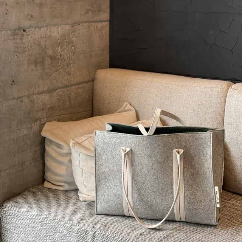 A gray felted wool shopping bag with beige handles placed on a sofa next to a cushion.