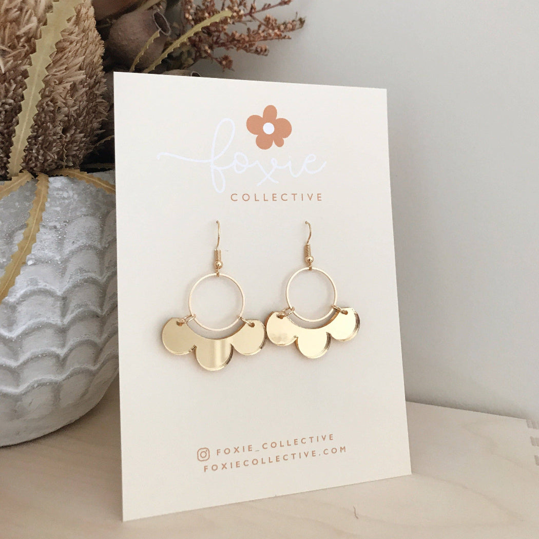 Gold-tone cloud-shaped dangle earrings displayed on a white card from Foxie Collective, set against a neutral background with dried floral decor.