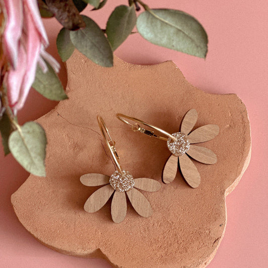 A pair of gold hoop earrings with flat wooden daisy flower petals and clear crystal centers, displayed on a terracotta flower-shaped stand against a pink background with green leaves and a pink flower as props.