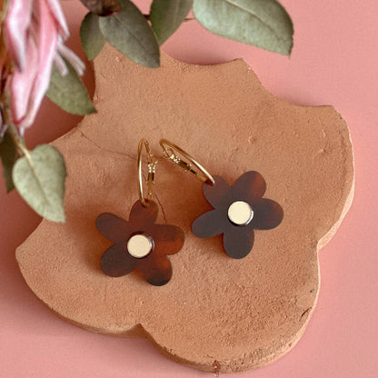 Flower-shaped earrings on a brown leaf with pink flowers and green leaves in the background