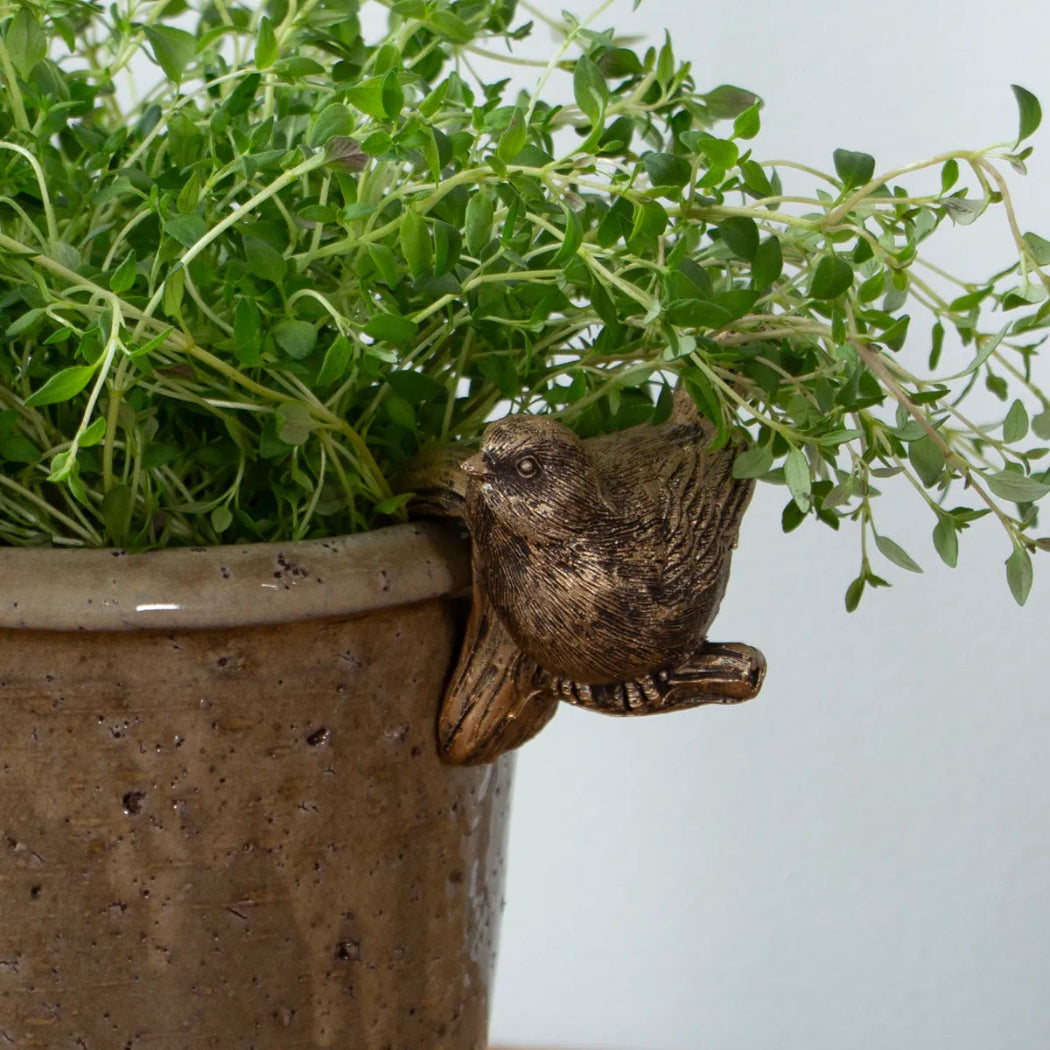 A small brown bird figurine perched on the rim of a speckled brown ceramic planter, with lush green leafy herbs growing out of the pot against a light gray wall.