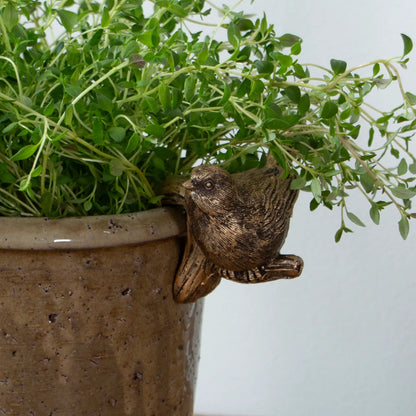 A small brown bird figurine perched on the rim of a speckled brown ceramic planter, with lush green leafy herbs growing out of the pot against a light gray wall.