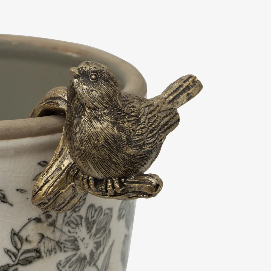 A close-up of a decorative metal bird figurine perched on the rim of a ceramic planter with black floral illustrations on a white background.