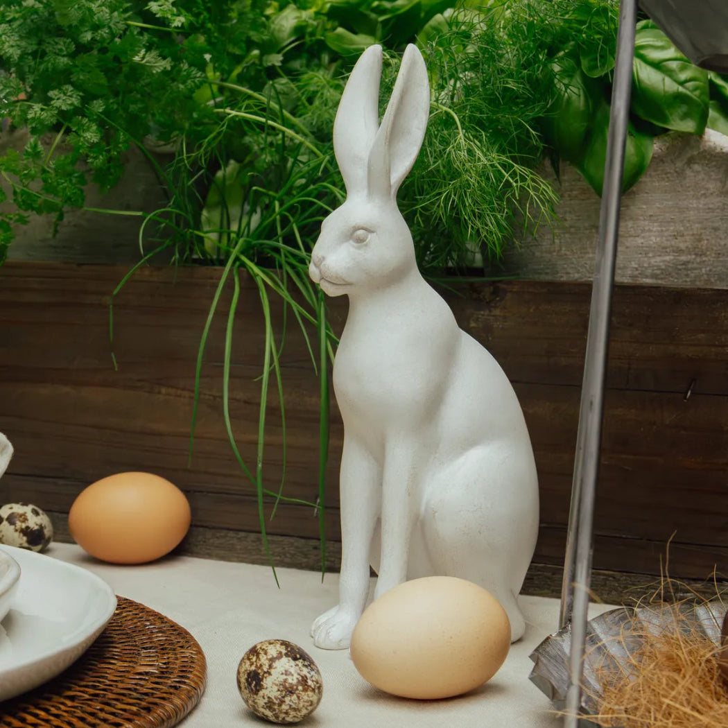 White ceramic rabbit figurine with Easter eggs on a wooden surface with greenery in the background