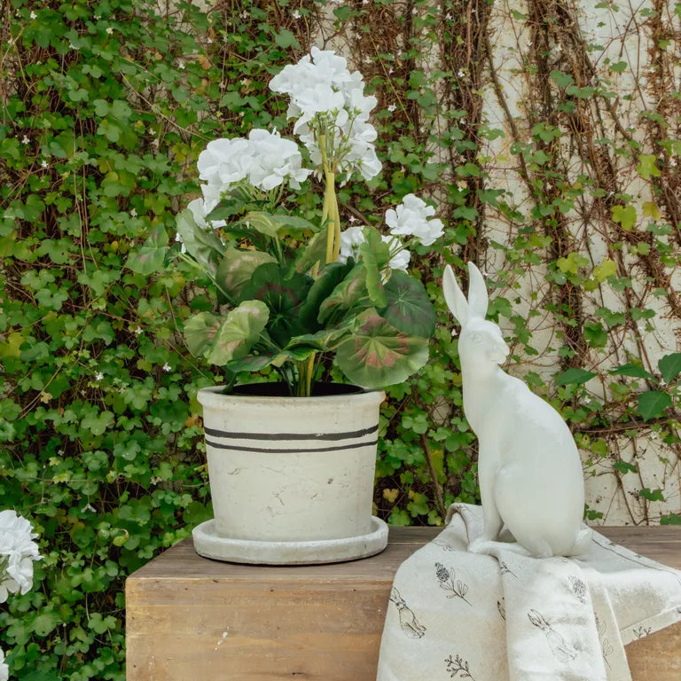 White rabbit figurine sitting next to a potted plant with white flowers on a wooden surface.