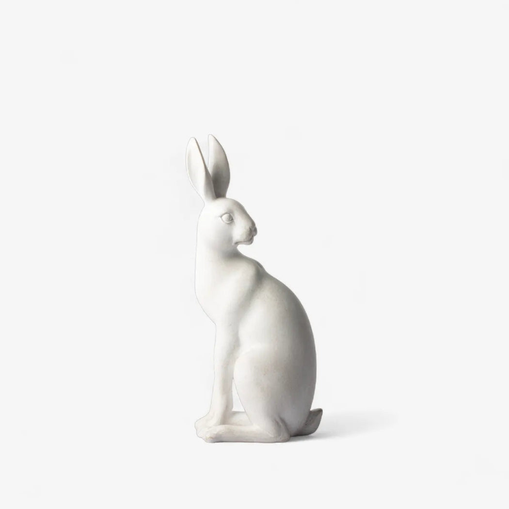 A minimalist white ceramic rabbit figurine in a seated profile position, photographed against a seamless, solid off-white background.