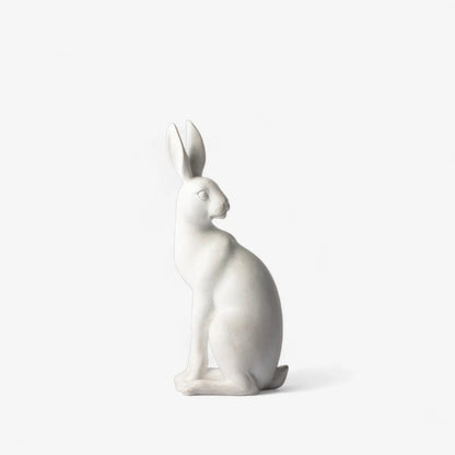 A minimalist white ceramic rabbit figurine in a seated profile position, photographed against a seamless, solid off-white background.