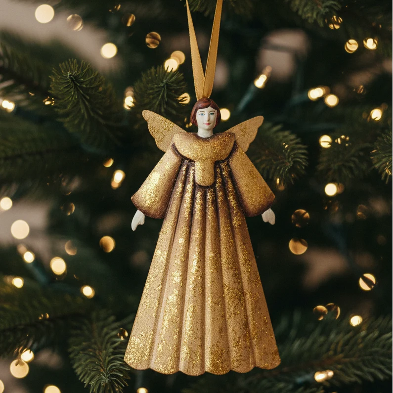 A gold glitter angel Christmas ornament hanging from a gold ribbon in front of a blurred Christmas tree with warm bokeh lights.