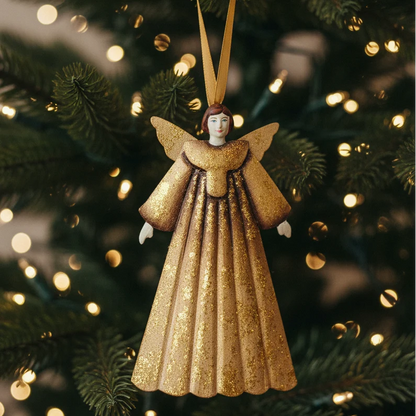 A gold glitter angel Christmas ornament hanging from a gold ribbon in front of a blurred Christmas tree with warm bokeh lights.