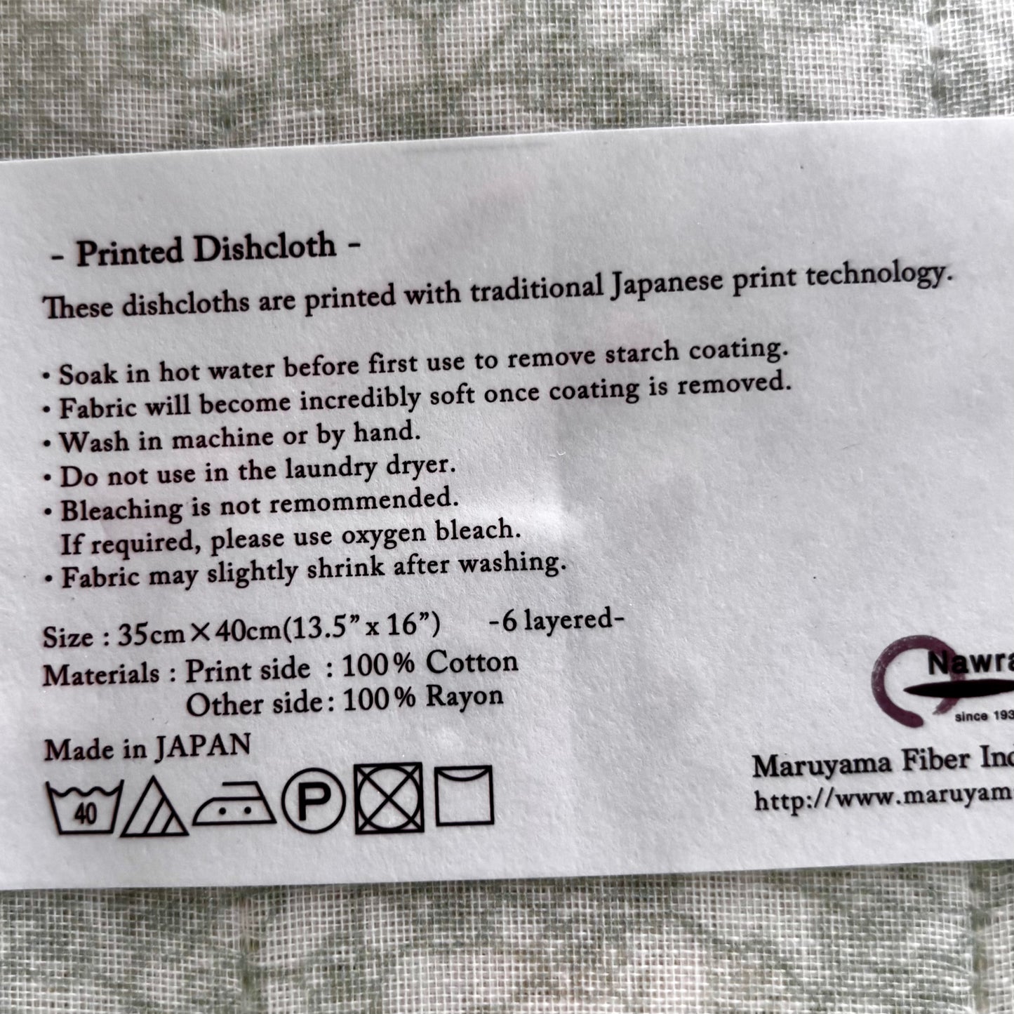 A printed white information card for a dishcloth, detailing care instructions, size (35cm x 40cm), material composition (100% cotton and 100% rayon), and manufacturing origin (Made in Japan). The card is placed on a textured gray fabric background.