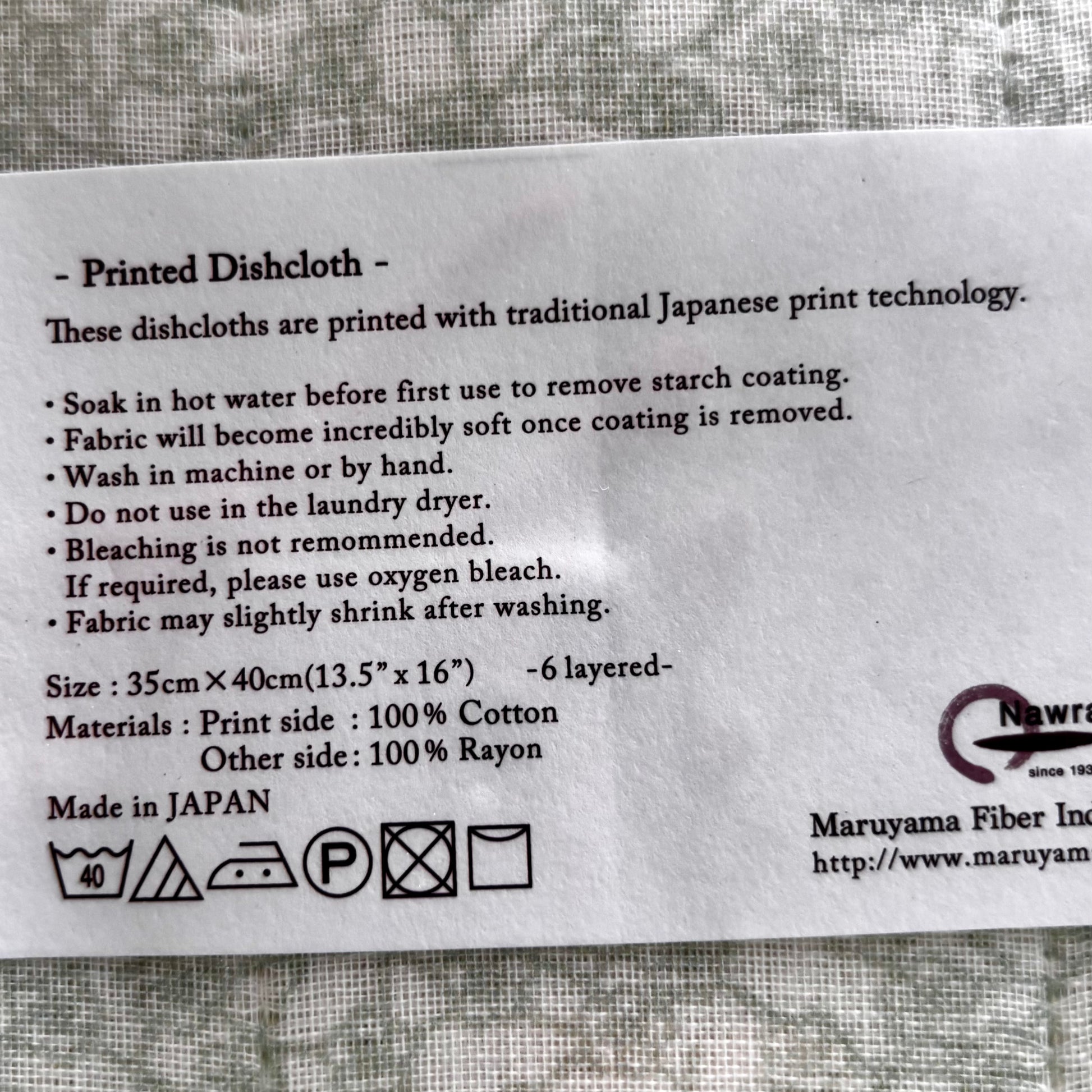 A printed white information card for a dishcloth, detailing care instructions, size (35cm x 40cm), material composition (100% cotton and 100% rayon), and manufacturing origin (Made in Japan). The card is placed on a textured gray fabric background.