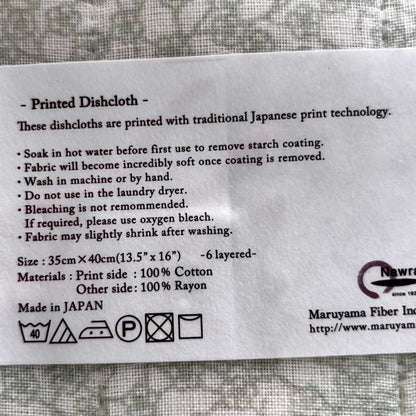 A printed white information card for a dishcloth, detailing care instructions, size (35cm x 40cm), material composition (100% cotton and 100% rayon), and manufacturing origin (Made in Japan). The card is placed on a textured gray fabric background.