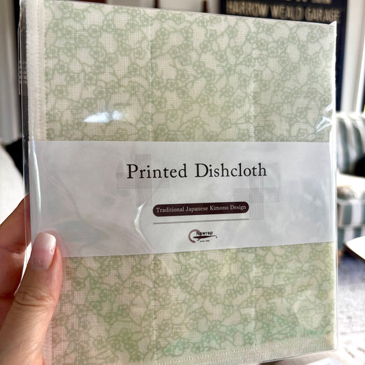 A hand holding a packaged dishcloth with a light green and cream Japanese kimono-style floral pattern. The product is wrapped in clear plastic with a white paper label that reads 'Printed Dishcloth' and shows the brand 'The Art of Life'.