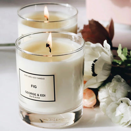 Two lit candles in glass containers with a 'Fig' label, surrounded by white flowers on a light background.
