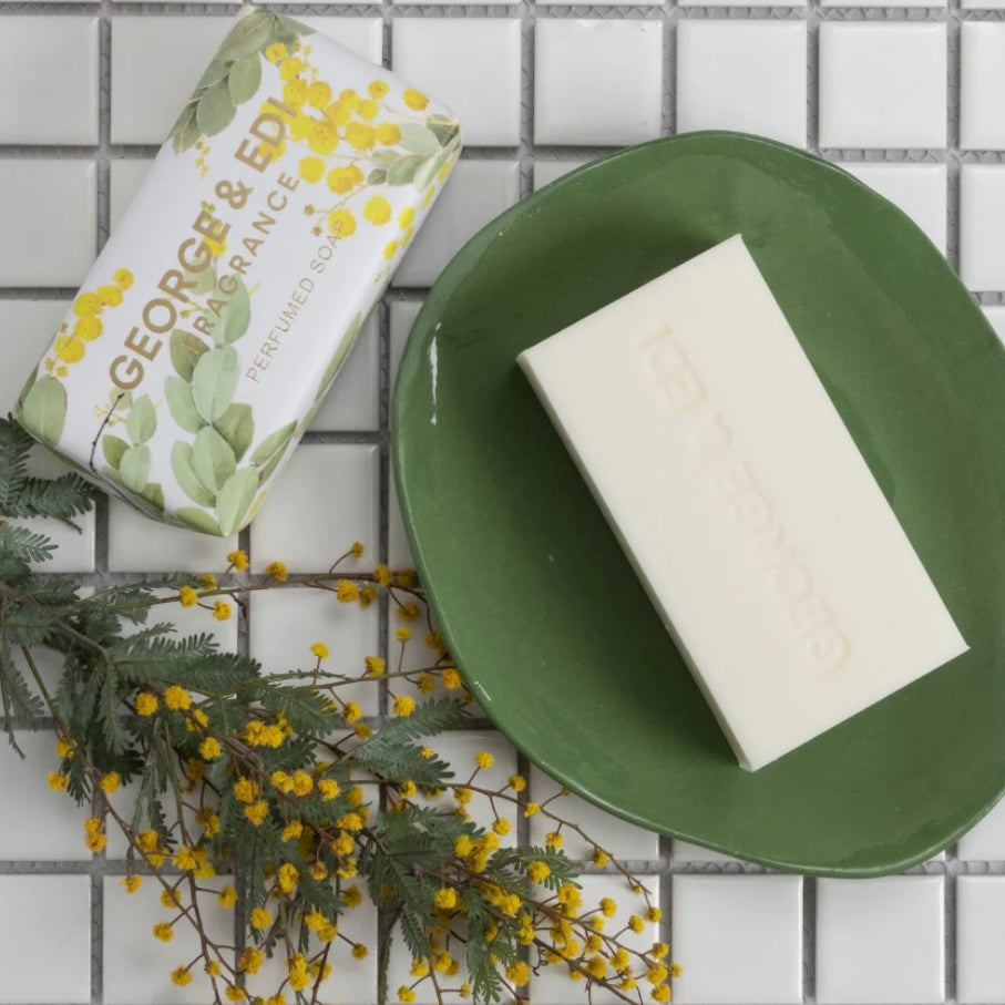Bar of soap on a green plate with a box labeled 'George & Edi' on a tiled background.