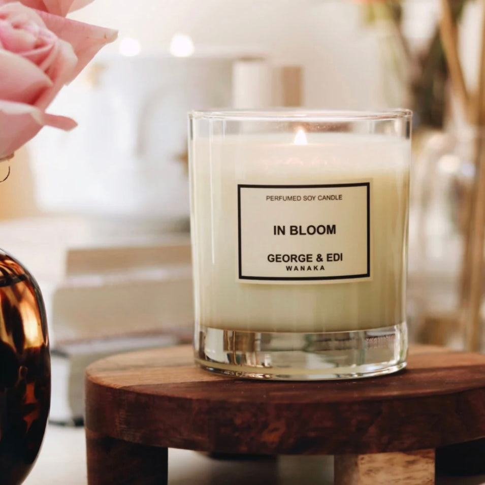 Candle labeled 'In Bloom' by George & Edi on a wooden stool with blurred background