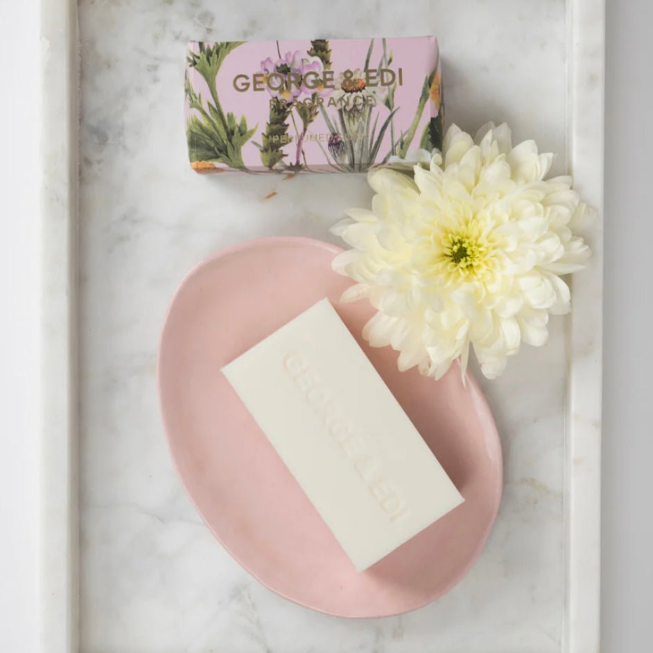 Bar of soap on a pink plate with a white flower and a box labeled 'Georges & Co.' on a marble surface.