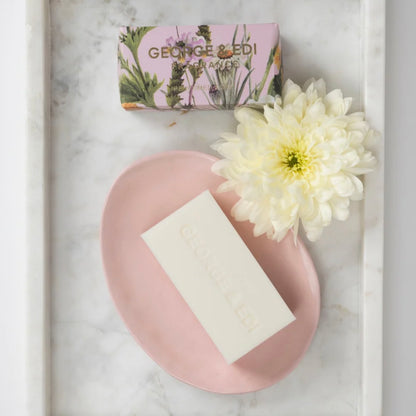 Bar of soap on a pink plate with a white flower and a box labeled 'Georges & Co.' on a marble surface.
